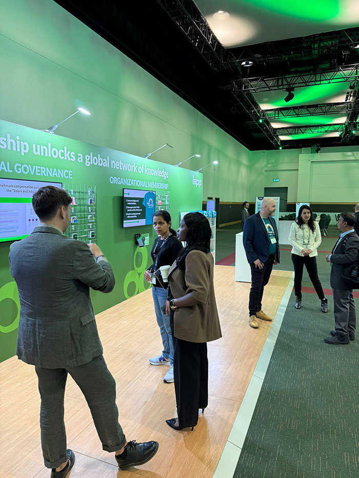 An exhibition area features a green wall display with informational panels and mounted screens labeled about organizational membership and global governance. Several attendees stand in small groups, conversing and looking at the displays under bright overhead lighting in a conference hall setting.