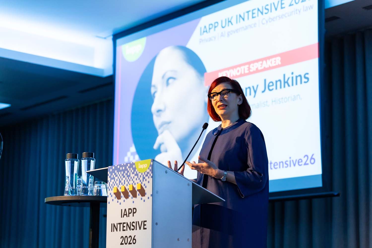 A speaker stands at a podium delivering a keynote while a large screen behind the stage displays conference branding and speaker information. Water glasses and IAPP signage are visible on the stage.