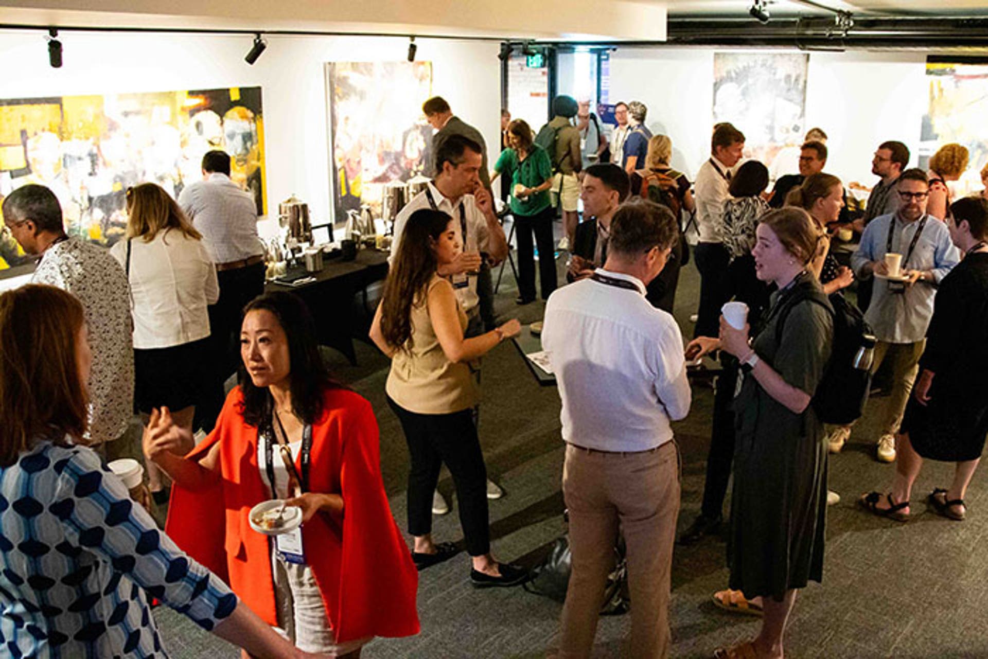 Networking event in a gallery space with people standing and talking near tables with coffee and refreshments. Colorful artwork displayed on the walls.