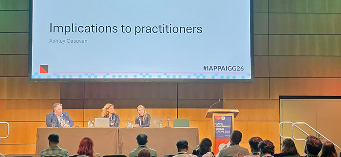 A panel of three speakers sits at a long table on a stage while a large screen above them displays the title Implications to practitioners and the name Ashley Casovan. Audience members are seated in rows facing the stage, and a podium stands to the right with event branding visible.