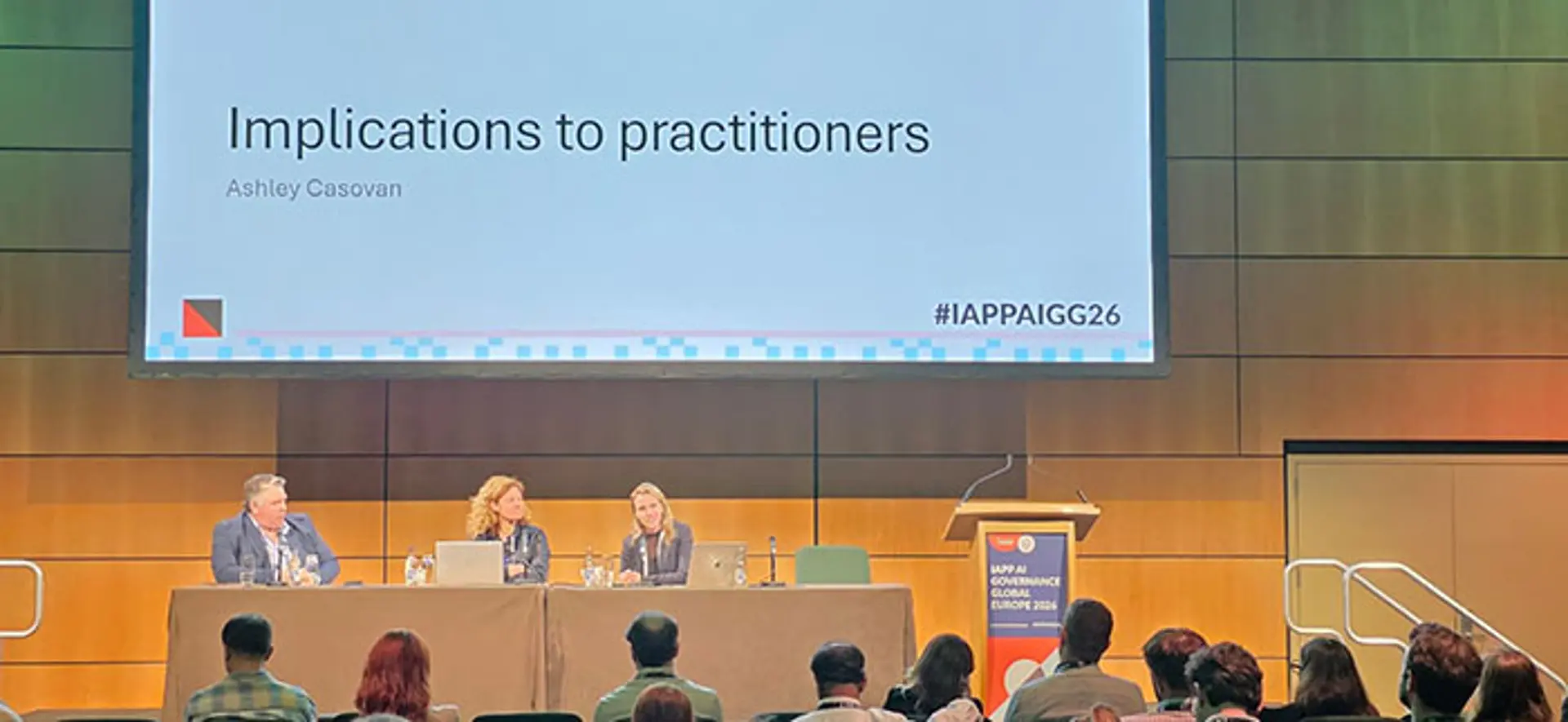 A panel of three speakers sits at a long table on a stage while a large screen above them displays the title Implications to practitioners and the name Ashley Casovan. Audience members are seated in rows facing the stage, and a podium stands to the right with event branding visible.