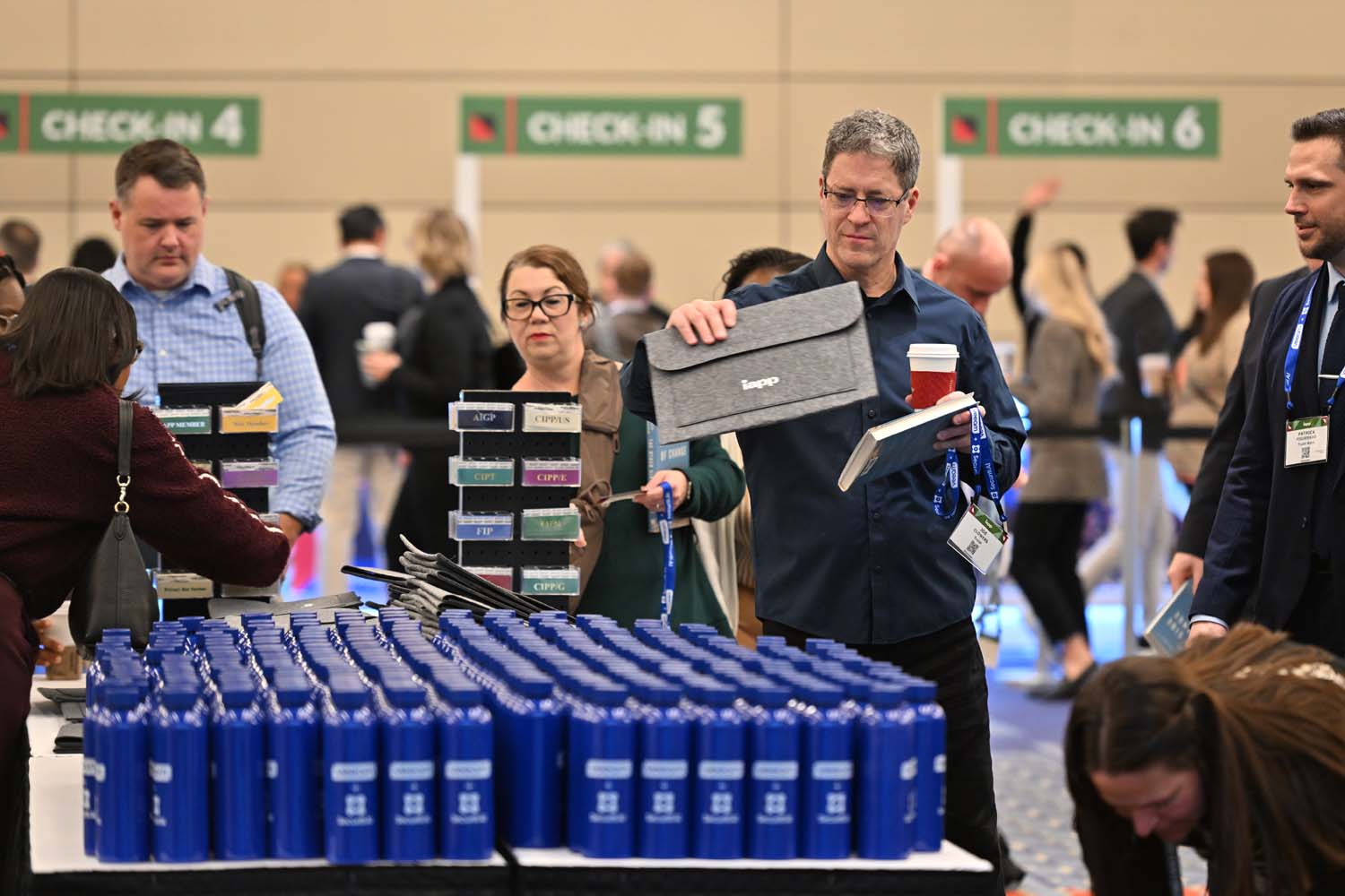 Attendees line up at a busy conference check-in area with rows of blue reusable water bottles in the foreground and credential displays and counters visible beneath overhead check-in signs.