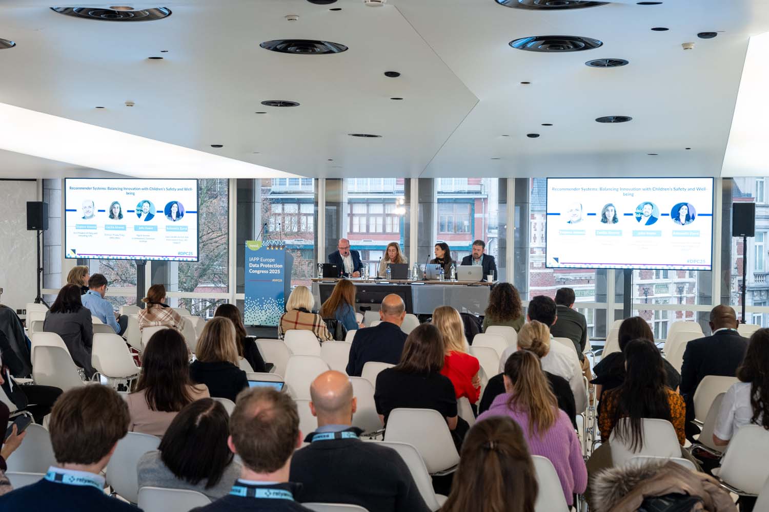 Conference session in a modern, bright room with large windows overlooking city buildings. Attendees are seated in rows facing a panel of four speakers at a long table. Two large screens display slides with headshots and text about “Responsible System Design.” A podium with an event sign reading “IAPP AI Governance Global Europe 2023” is positioned near the panel.