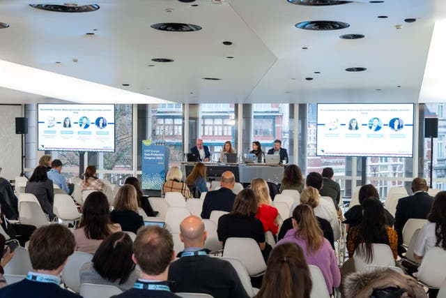 Conference session in a modern, bright room with large windows overlooking city buildings. Attendees are seated in rows facing a panel of four speakers at a long table. Two large screens display slides with headshots and text about “Responsible System Design.” A podium with an event sign reading “IAPP AI Governance Global Europe 2023” is positioned near the panel.