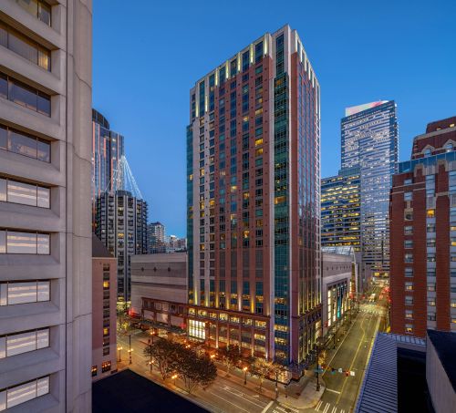 A reflective glass skyscraper rises above neighboring towers under a bright sky, its mirrored facade catching clouds and sunlight while the city stretches out in layered depth below.
