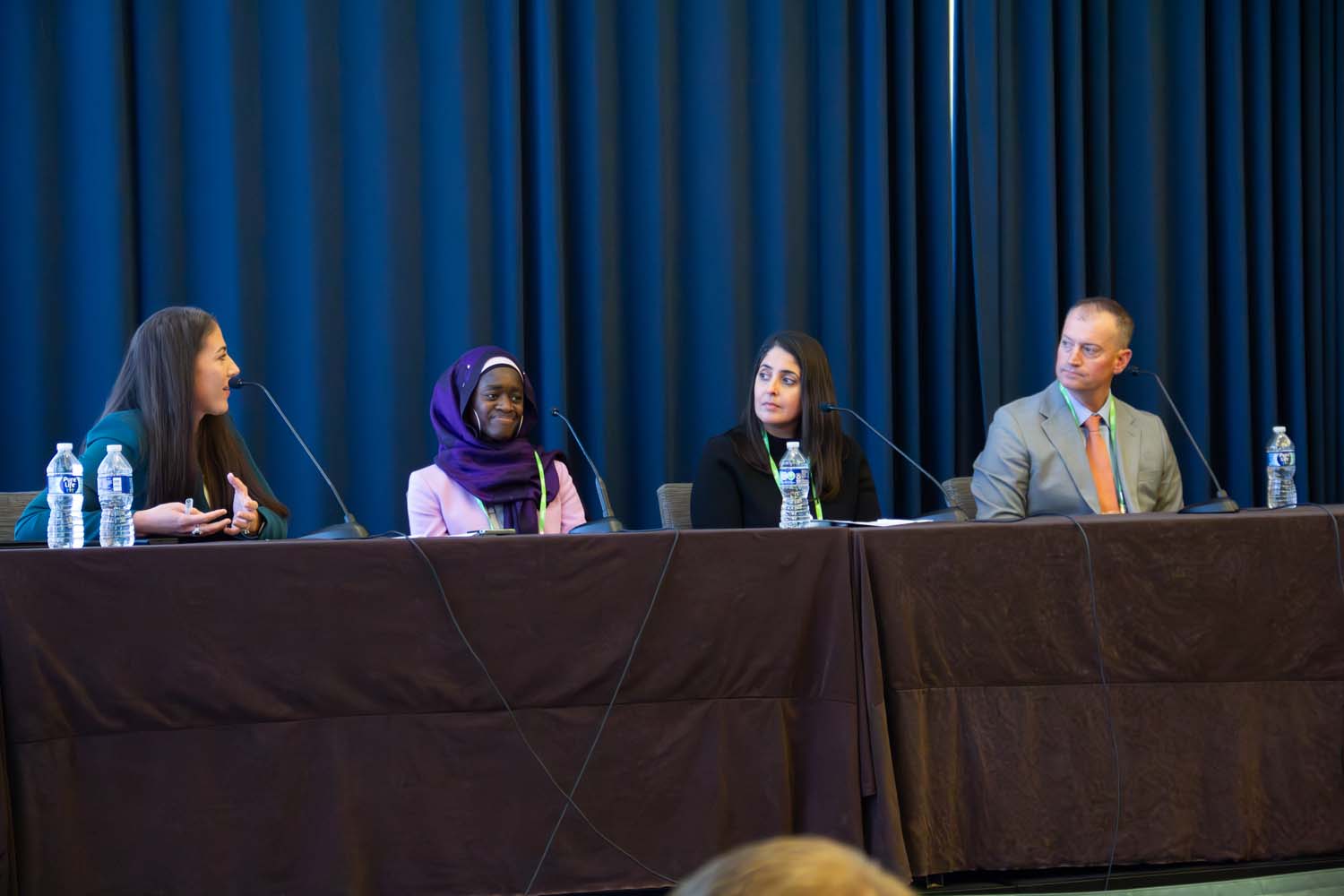 A full conference room is shown from the back, with attendees seated in rows facing a stage where a panel discussion is displayed on dual screens at the front of the room.