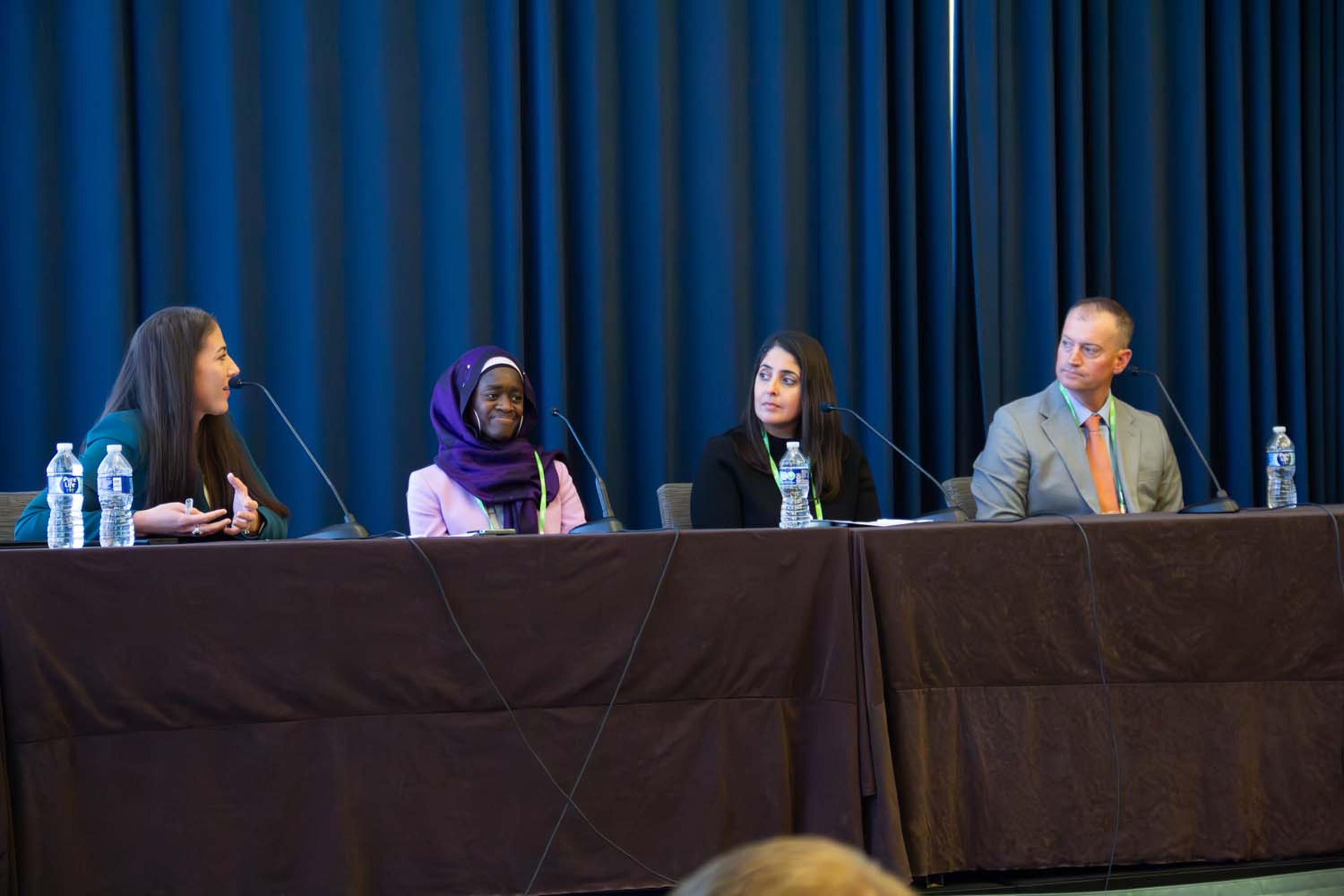 A full conference room is shown from the back, with attendees seated in rows facing a stage where a panel discussion is displayed on dual screens at the front of the room.