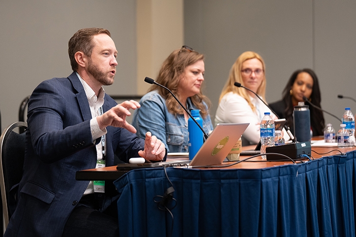Panel discussion with four people seated at a long table covered in a blue tablecloth. Microphones, laptops, and water bottles are on the table. The setting appears to be a conference room with neutral walls.