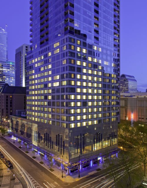 A glass-wrapped high-rise glows with warm yellow light at dusk, its stacked floors and blue-lit base rising above an active city intersection streaked with passing traffic.