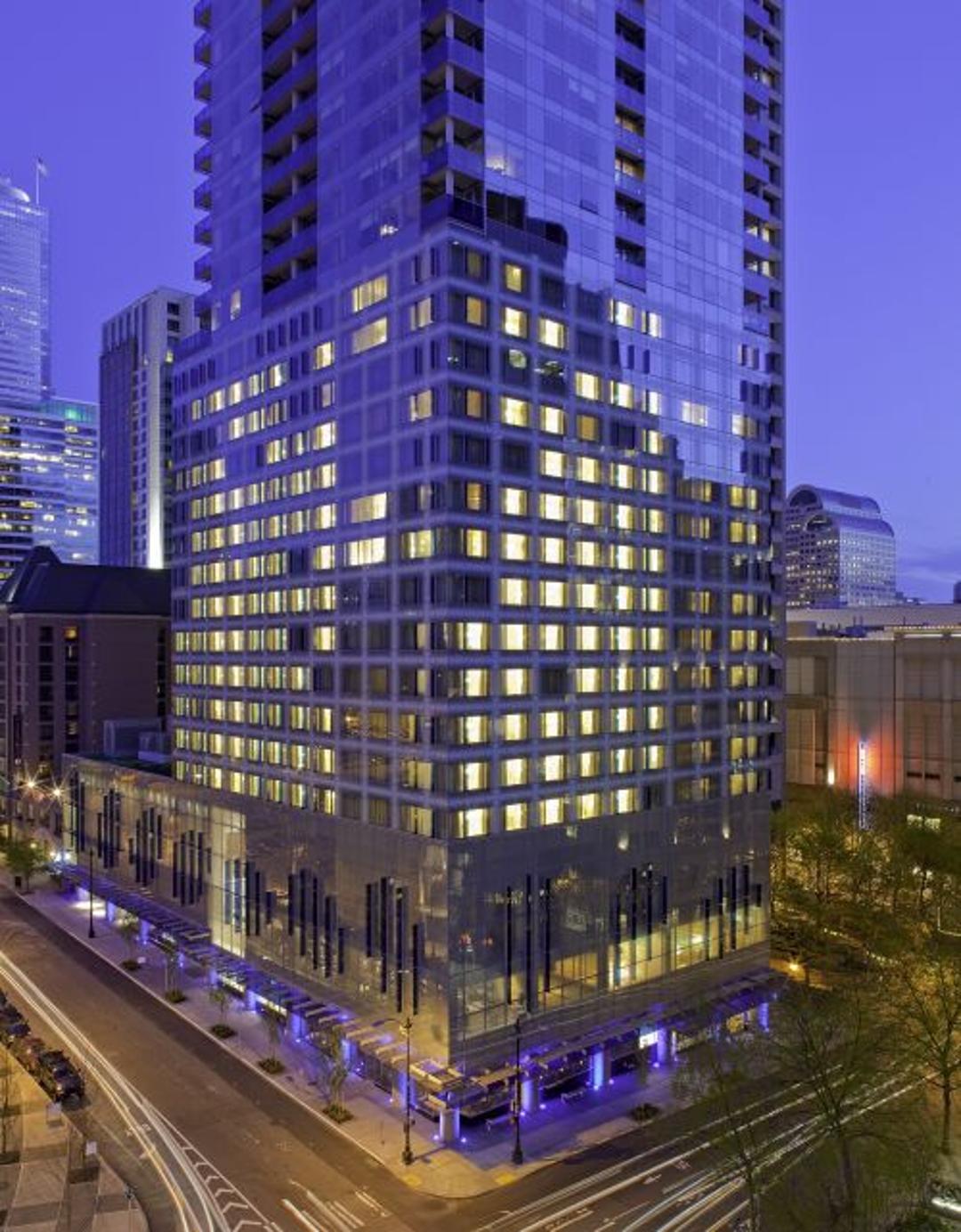 A glass-wrapped high-rise glows with warm yellow light at dusk, its stacked floors and blue-lit base rising above an active city intersection streaked with passing traffic.