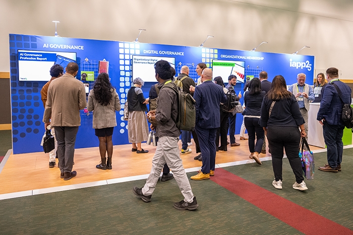 Exhibit area with multiple attendees standing and walking in front of a bright blue display wall featuring three sections labeled “AI Governance,” “Digital Governance,” and “Organizational Membership.” Each section includes screens and informational graphics. The floor has a red carpet strip, and the event branding “iapp25” is visible on the far right.