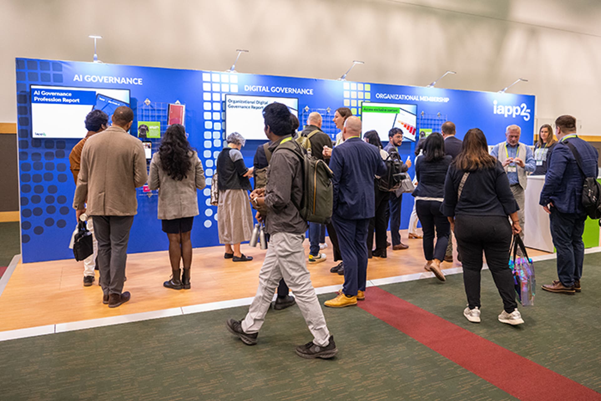 Exhibit area with multiple attendees standing and walking in front of a bright blue display wall featuring three sections labeled “AI Governance,” “Digital Governance,” and “Organizational Membership.” Each section includes screens and informational graphics. The floor has a red carpet strip, and the event branding “iapp25” is visible on the far right.