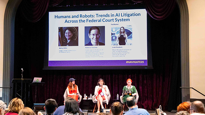 Panel discussion in a venue with a purple curtain backdrop. A large screen shows the session title: “Humans and Robots: Trends in AI Litigation Across the Federal Court System.”