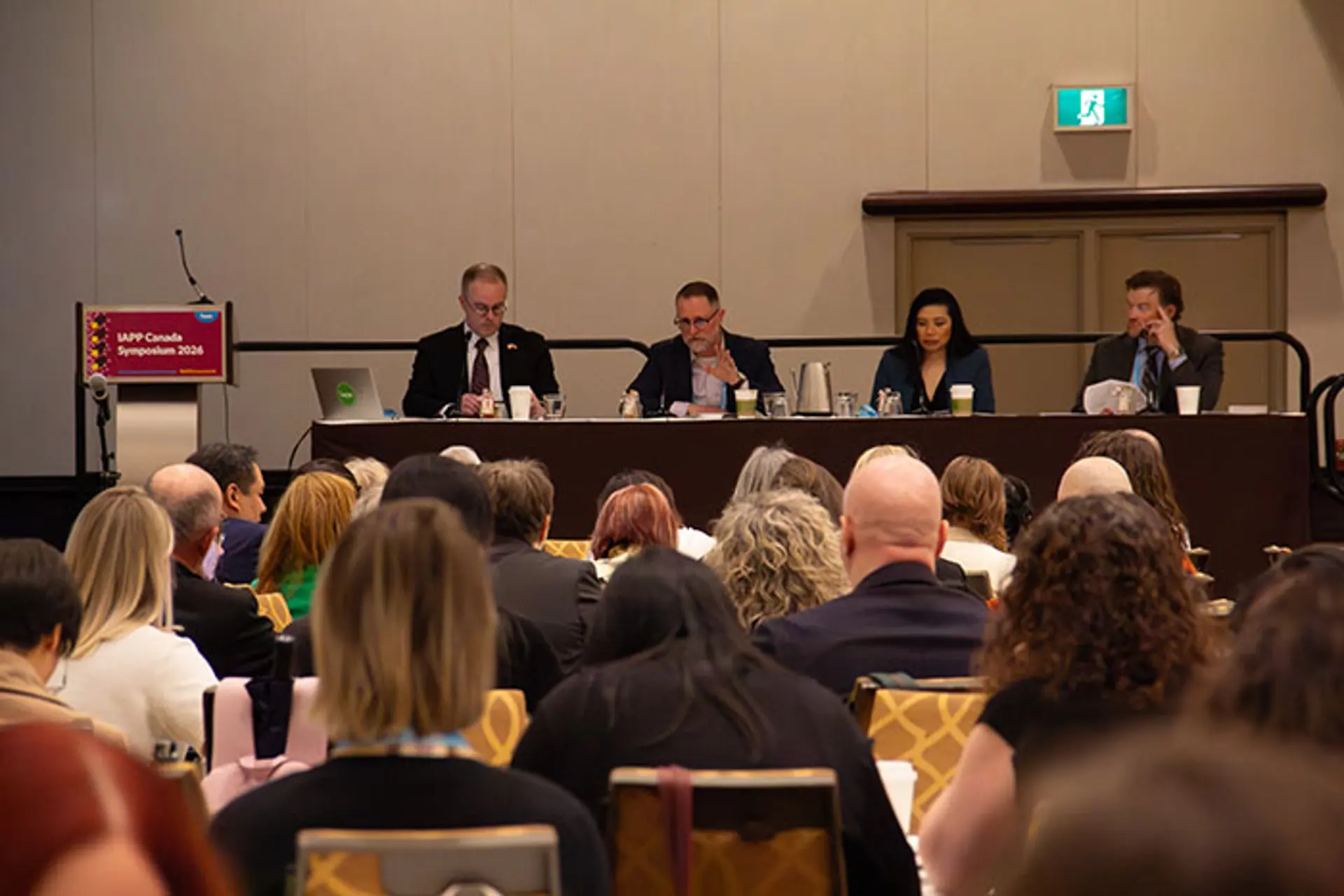 Panel of speakers seated behind a long table in a conference room addressing an audience, with attendees visible from behind and event signage reading “IAPP Canada Symposium 2026” on a podium.