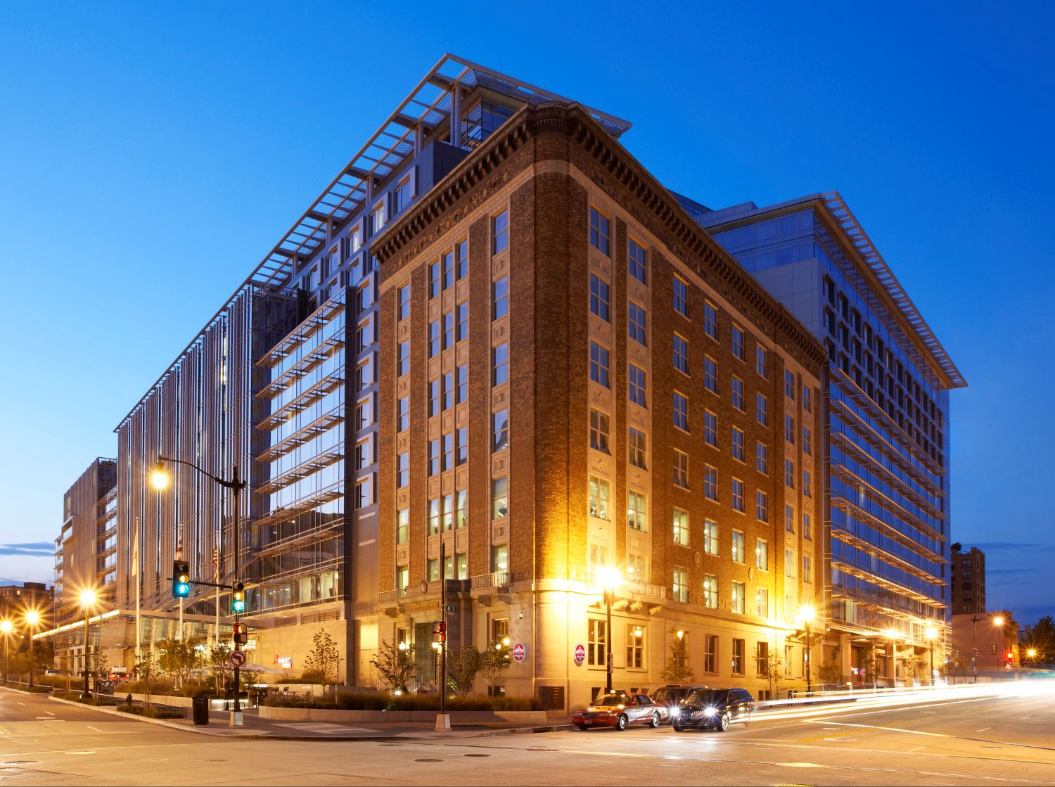 A large multi‑story hotel building at a city intersection combines historic brick architecture with modern glass and steel additions. Streetlights and passing cars illuminate the scene at dusk against a clear blue sky.