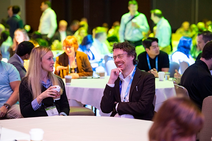 Two attendees seated at a round table during a breakout session, holding coffee cups. Other participants are visible in the background under green and yellow lighting.
