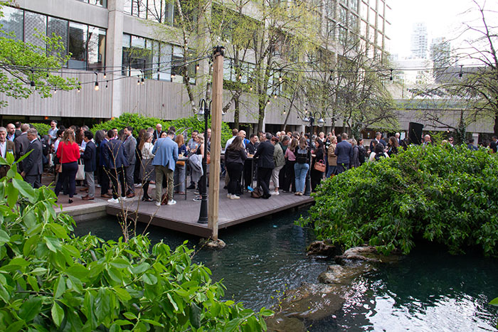 Crowd of attendees mingling on an outdoor platform next to water and landscaped plants, with high-top tables and surrounding trees in a courtyard setting.