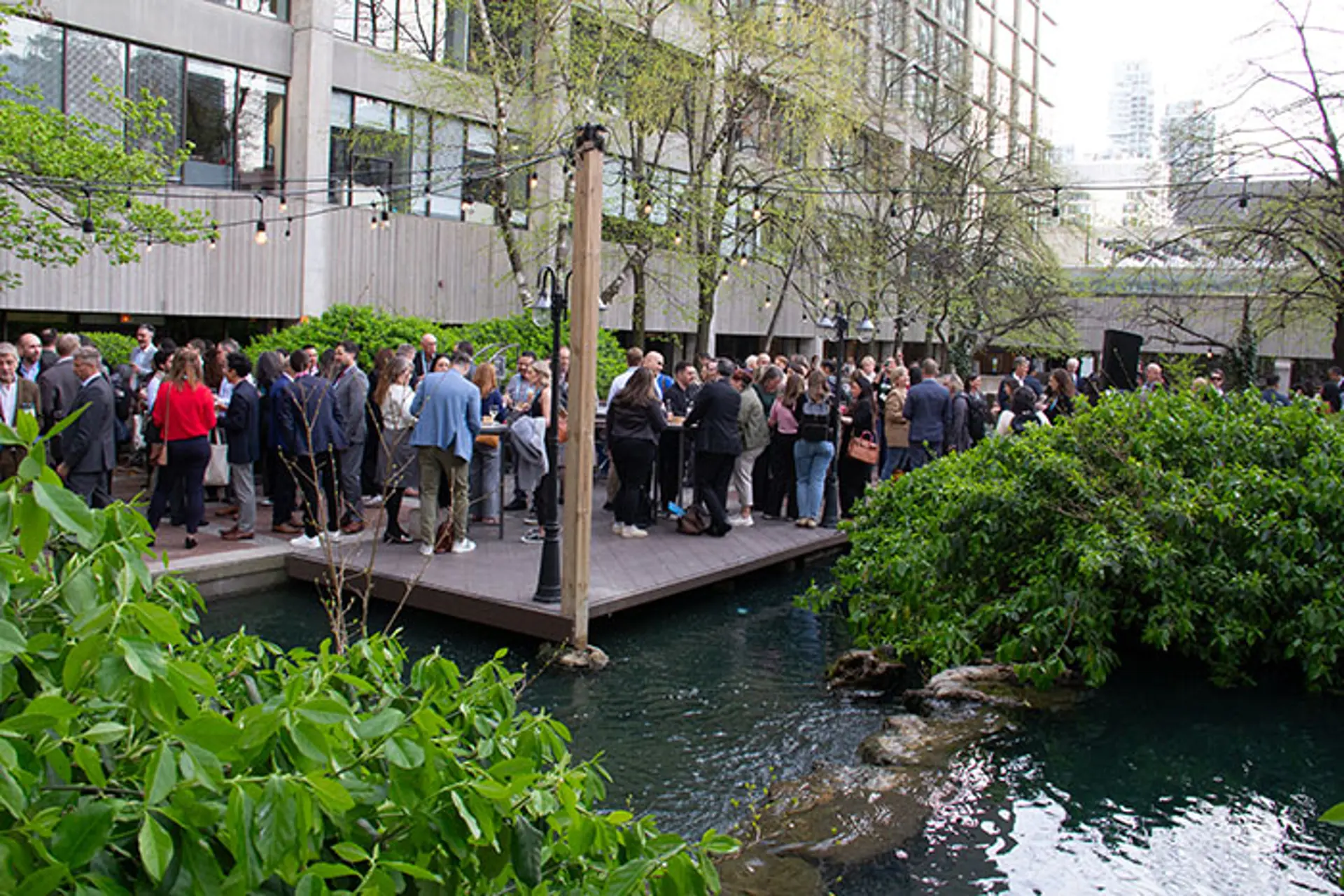 Crowd of attendees mingling on an outdoor platform next to water and landscaped plants, with high-top tables and surrounding trees in a courtyard setting.