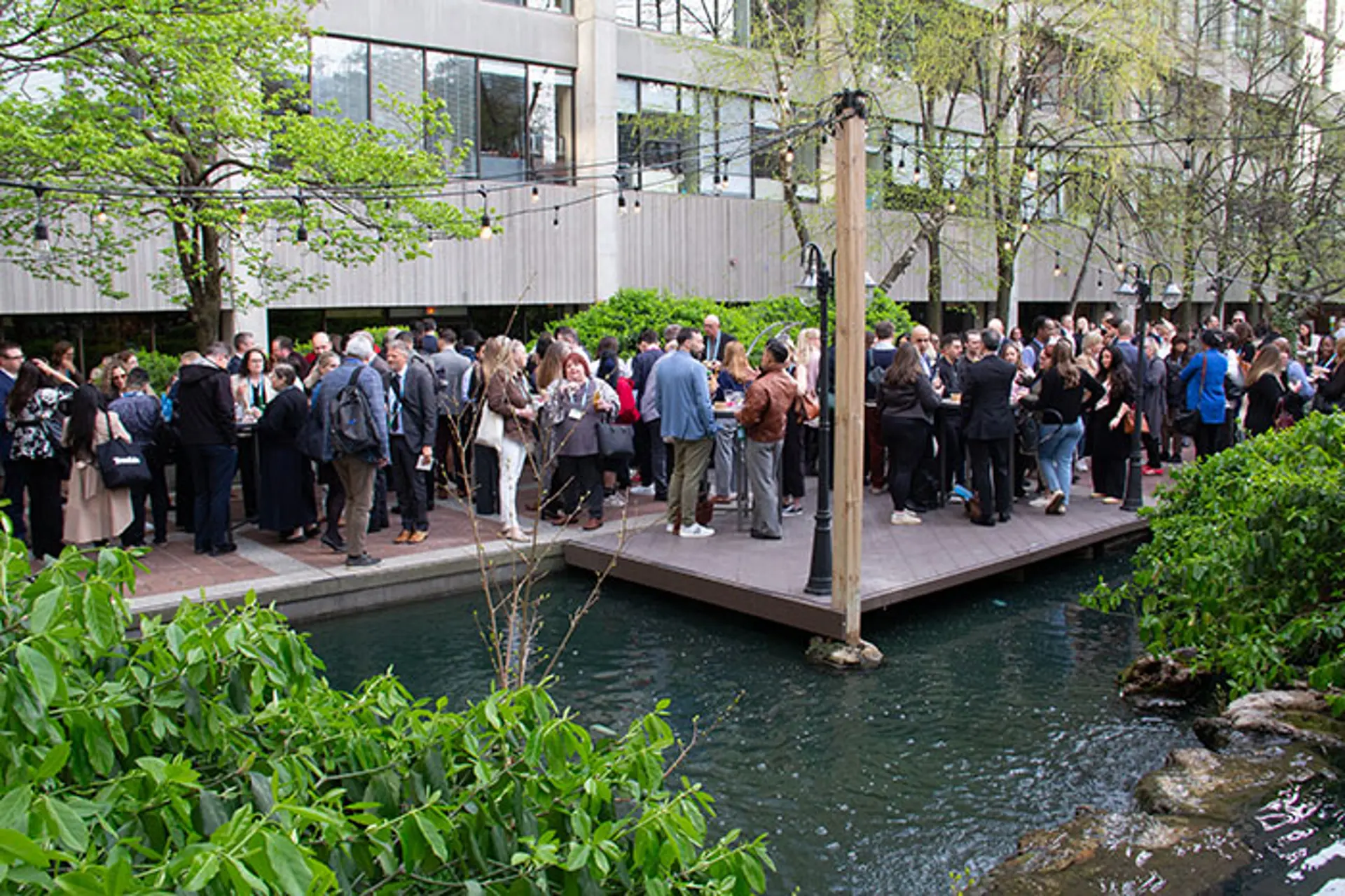 Large group of conference attendees gathered outdoors on a wooden deck beside a pond, networking under string lights with greenery and a multi-story building in the background.