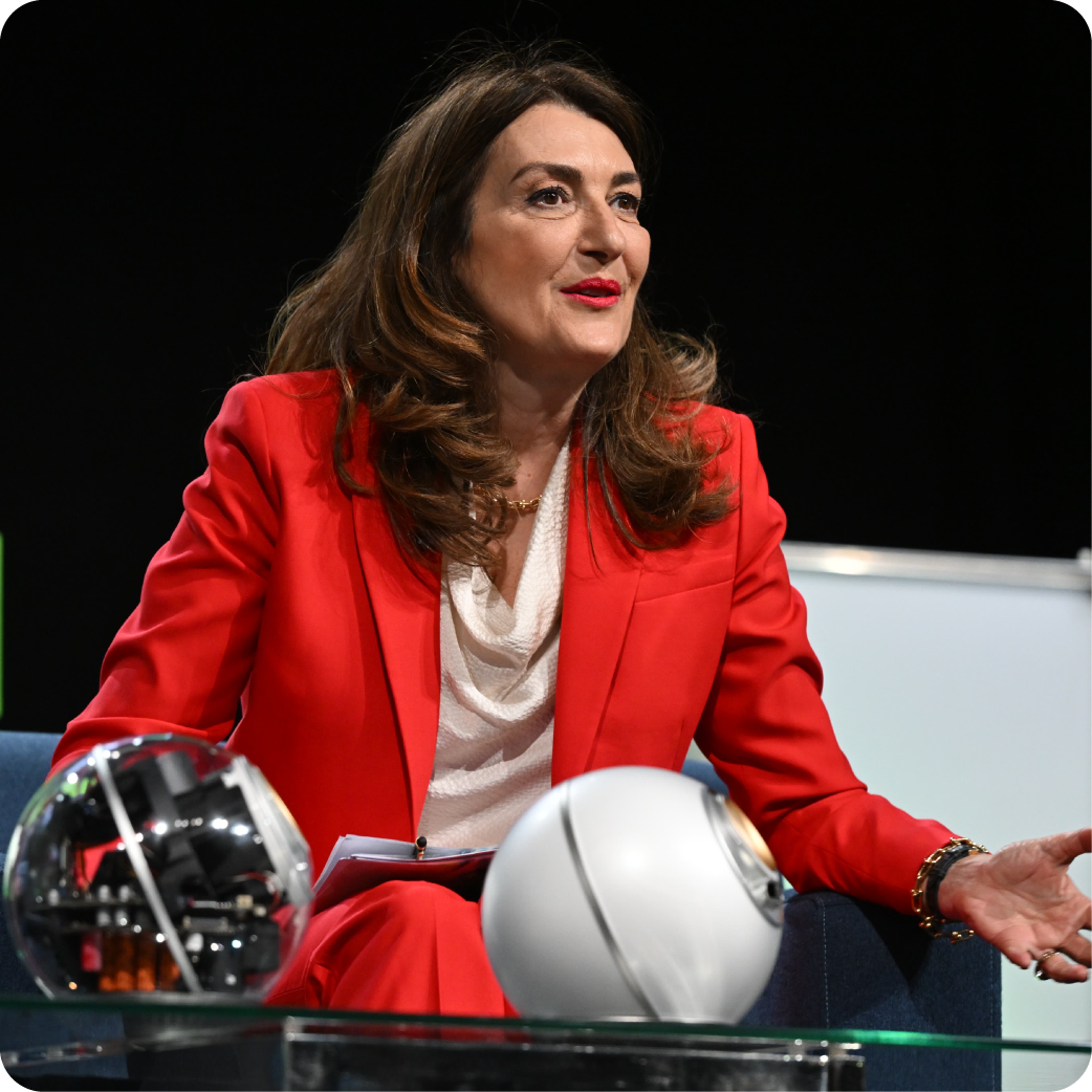 A woman in a red jacket sitting down and speaking.