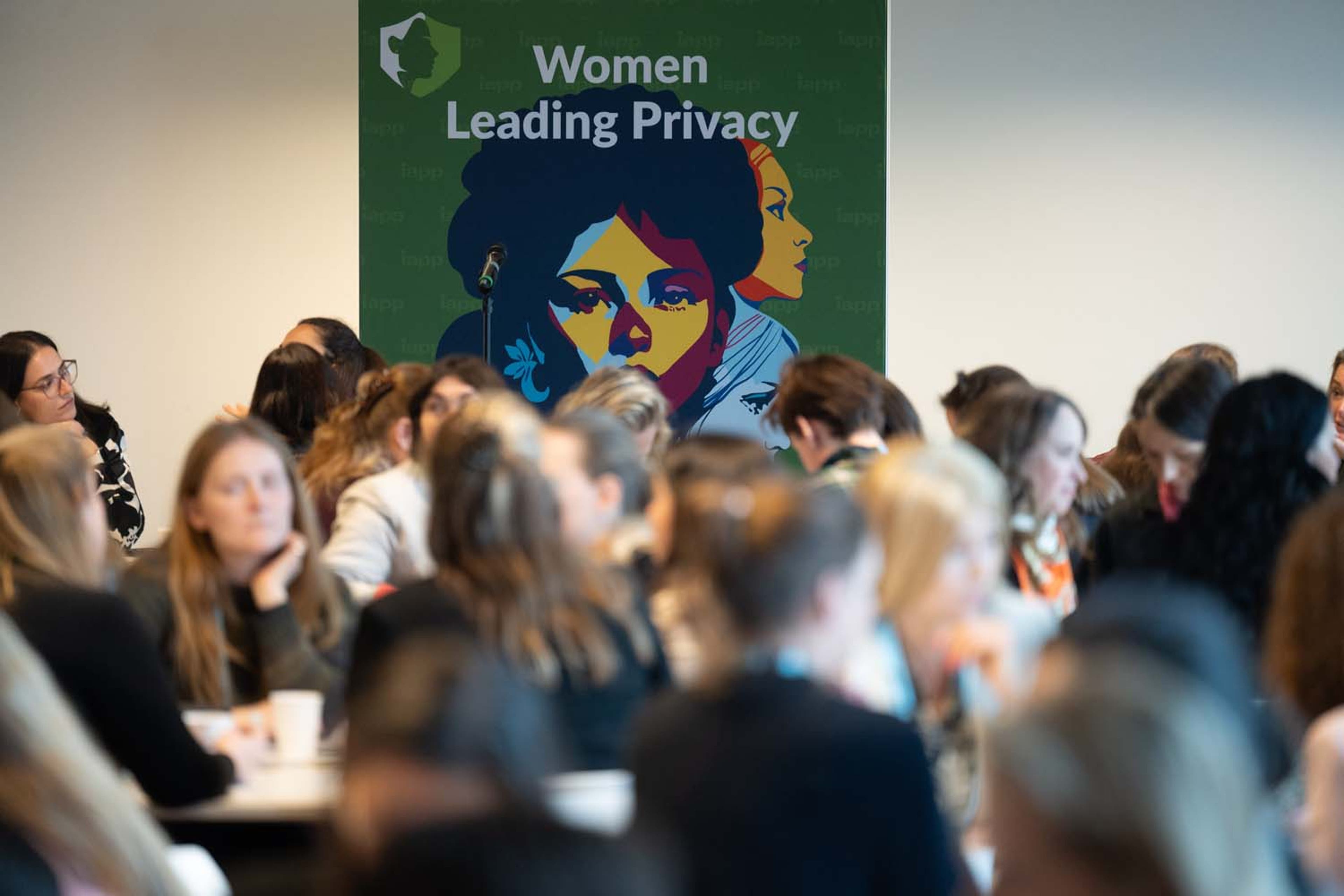 Crowded room with delegates seated at tables during a “Women Leading Privacy” event. A green banner with the event title is visible in the background.