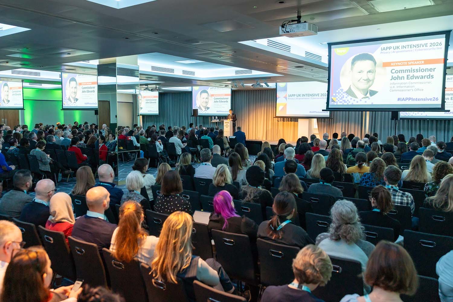 A large audience fills a conference hall facing a stage with a podium and multiple presentation screens. Event branding and keynote session visuals are displayed prominently.