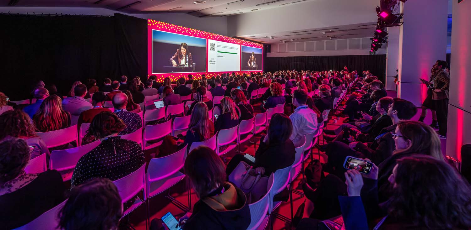 Packed conference session with rows of delegates seated facing a brightly lit stage. A large screen displays a speaker and presentation slides under pink lighting.