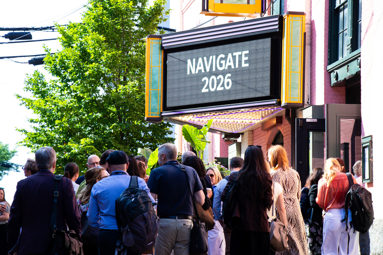 A crowd of conference attendees gathers on a sunlit city sidewalk beneath a vintage theater marquee displaying the text “NAVIGATE 2026,” with leafy green trees and historic building facades framing the entrance to the event.