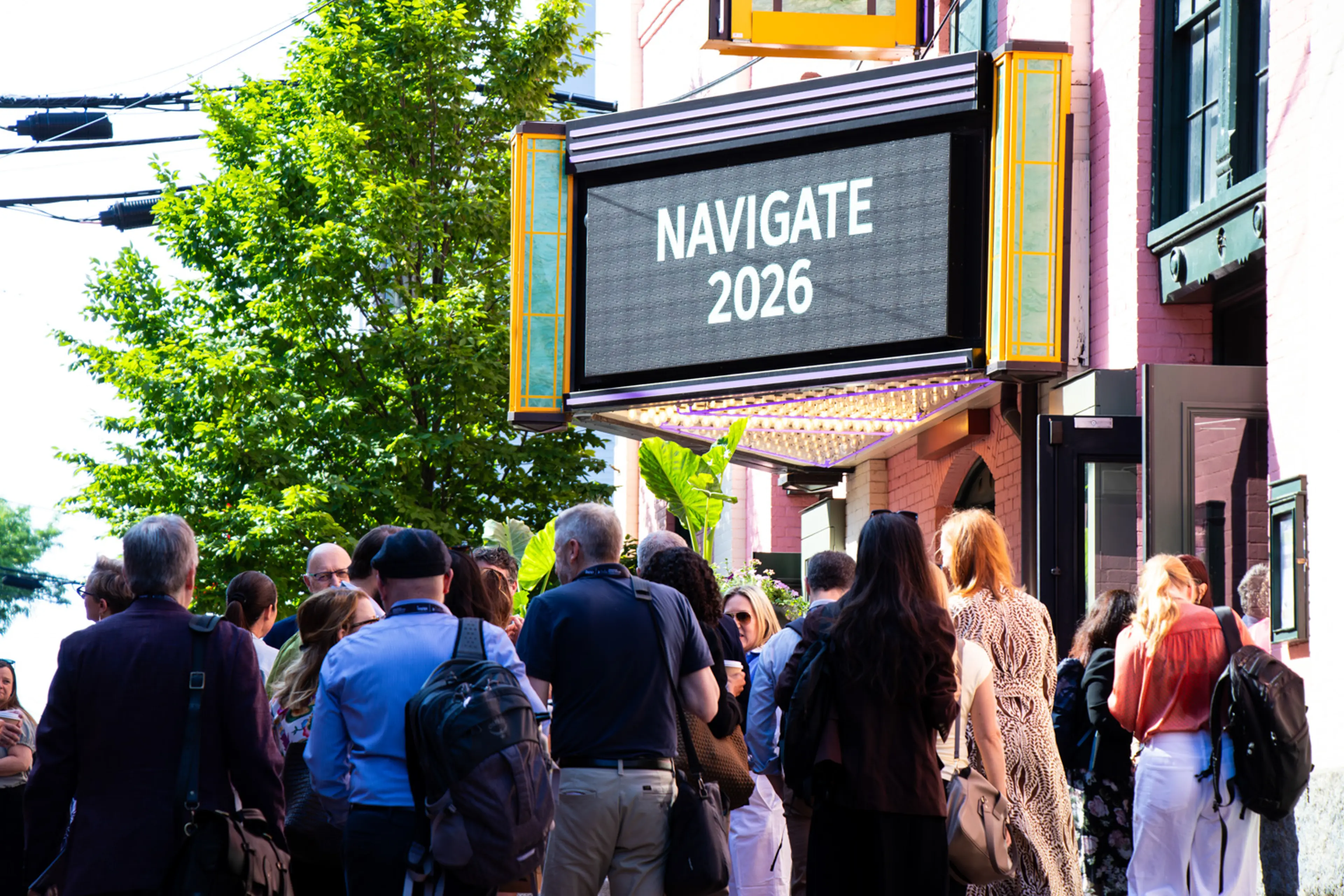 A crowd of conference attendees gathers on a sunlit city sidewalk beneath a vintage theater marquee displaying the text “NAVIGATE 2026,” with leafy green trees and historic building facades framing the entrance to the event.