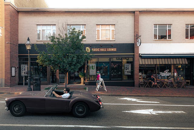 Street view of a beige two-story building with a black sign reading “Music Hall Lounge.” A tree stands in front of the entrance, and a person walks on the brick sidewalk carrying a sandwich board sign. To the right, an outdoor café has tables and chairs with people seated. In the foreground, a vintage dark red convertible car is driving past on the road with white directional arrows painted on the pavement.