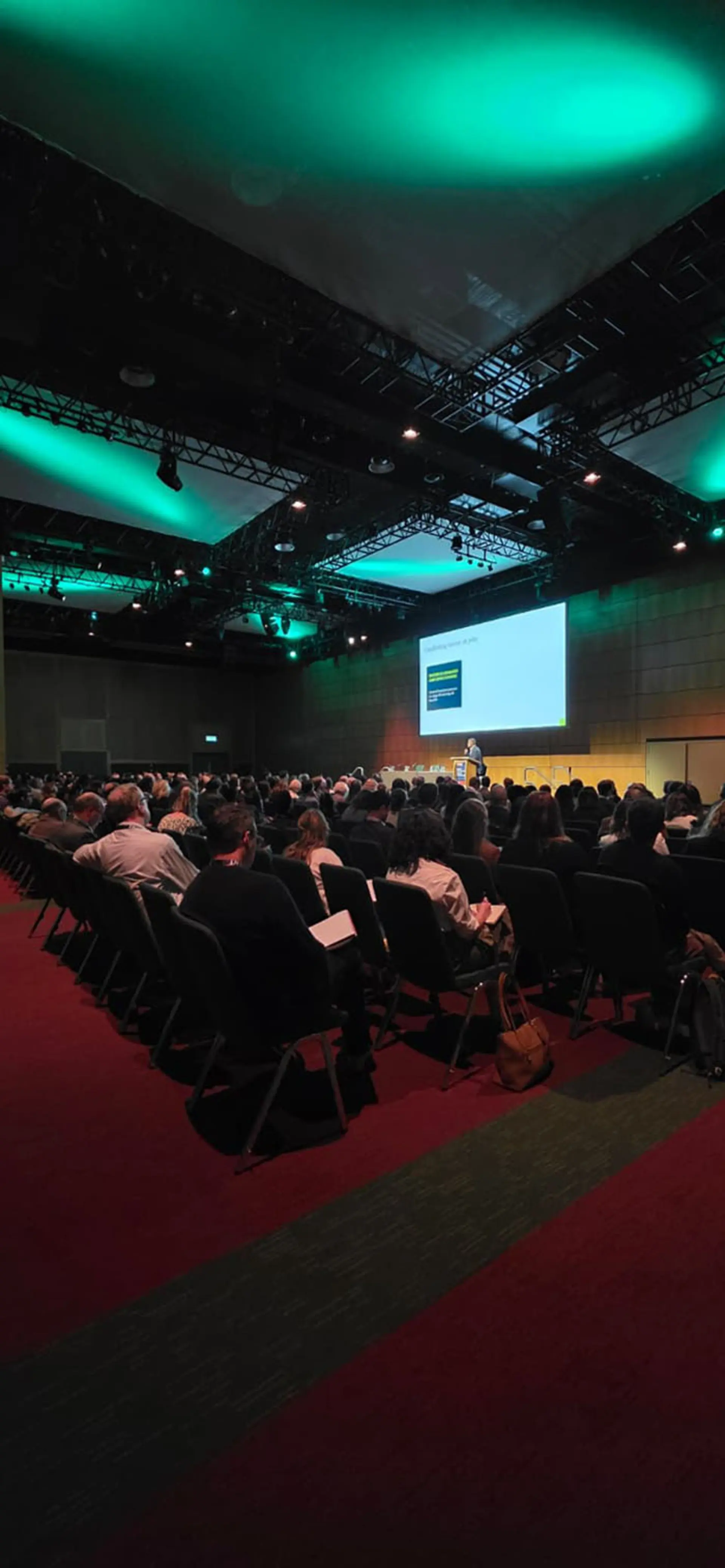 A large conference hall shows rows of seated attendees facing a stage where a presenter stands near a podium beside a large projection screen displaying a slide. Overhead rigging and lighting cast green and white illumination across the ceiling, and the room is arranged in a theater-style layout with a central aisle and red carpeting.