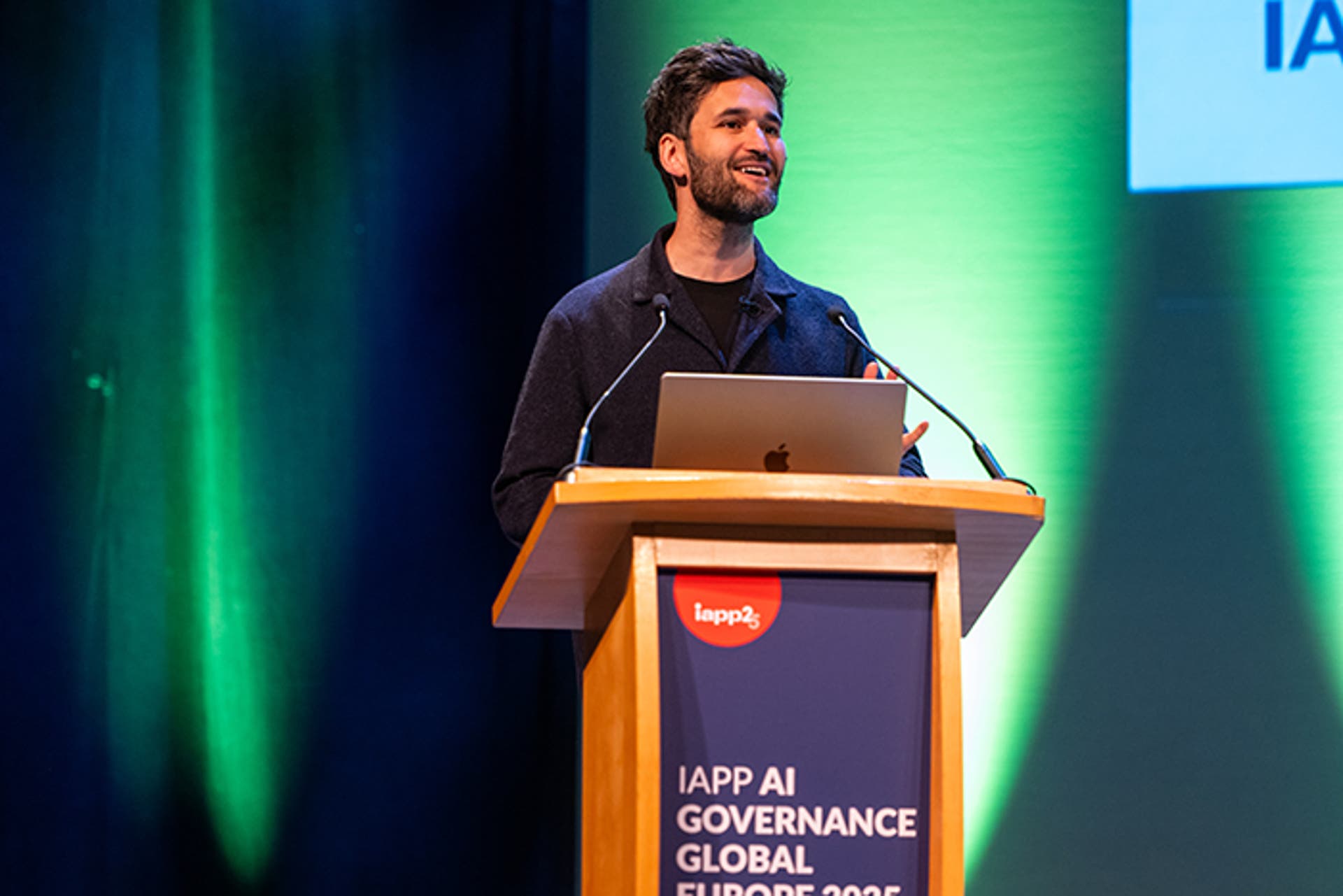 Speaker presenting at a podium with a laptop on stage. Podium signage reads “IAPP AI Governance Global Europe 2025.” Green lighting in the background.