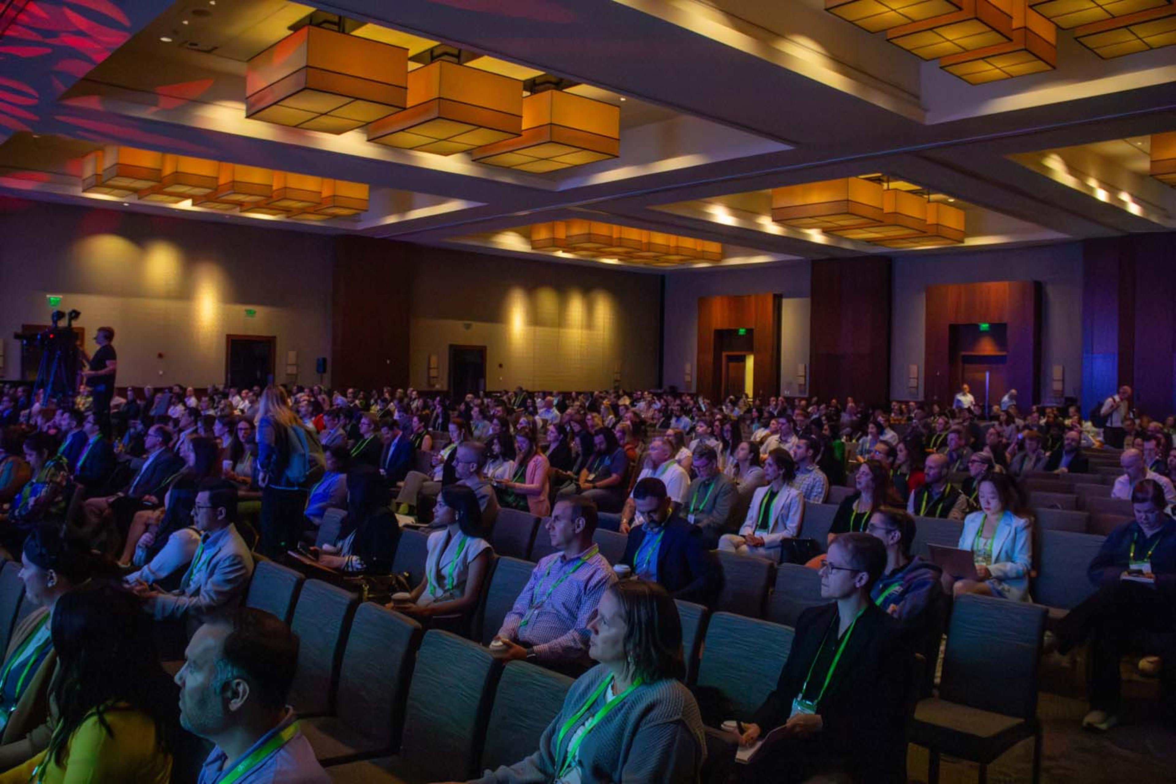 A wide view of a packed conference hall shows hundreds of attendees seated beneath geometric ceiling lights, focused on the stage during an IAPP AI Governance plenary session.