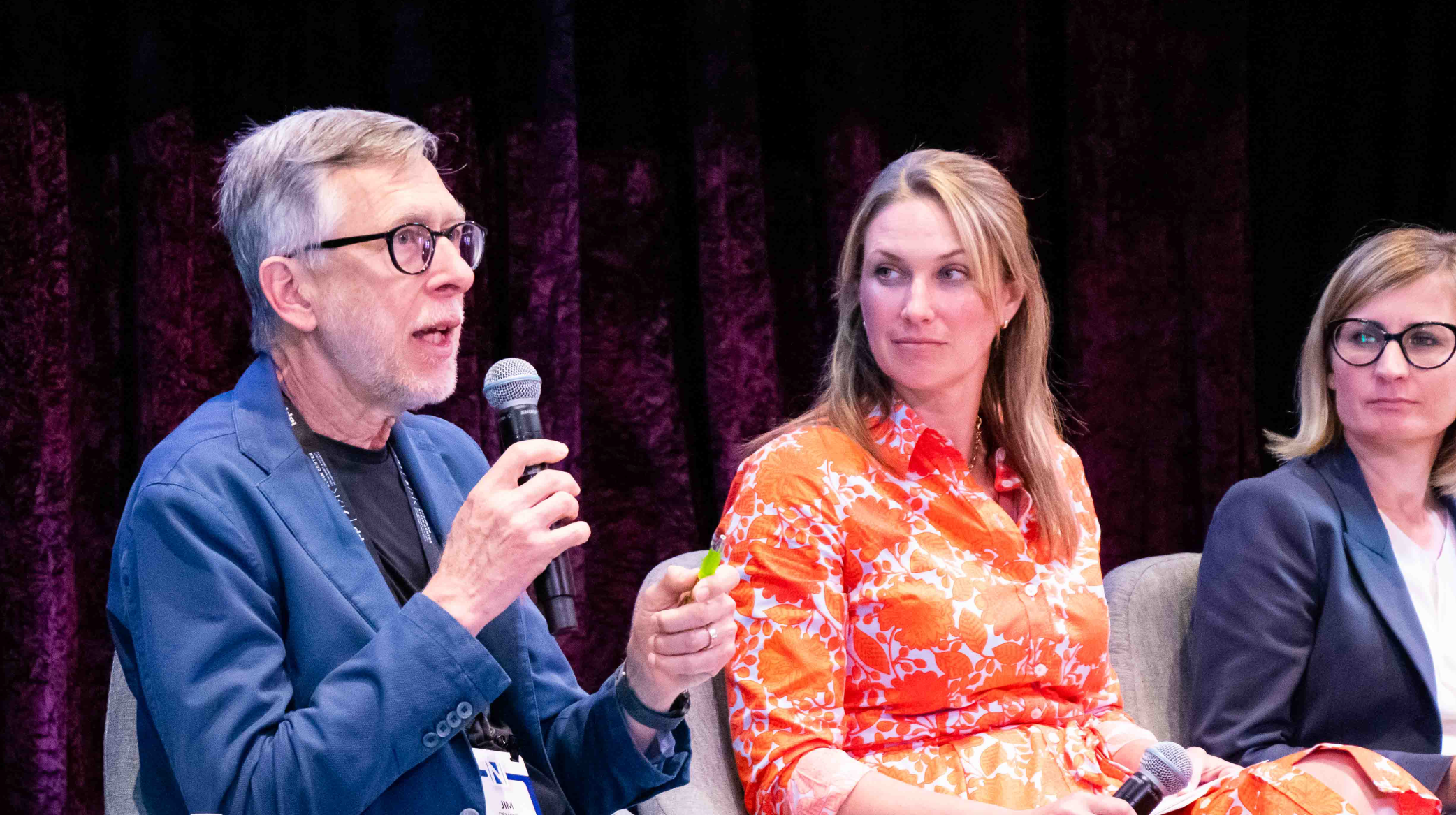 Three people on a stage taking part in a panel discussion. In the left foreground is a man in glasses speaking into a microphone; to his right are two women on the panel.