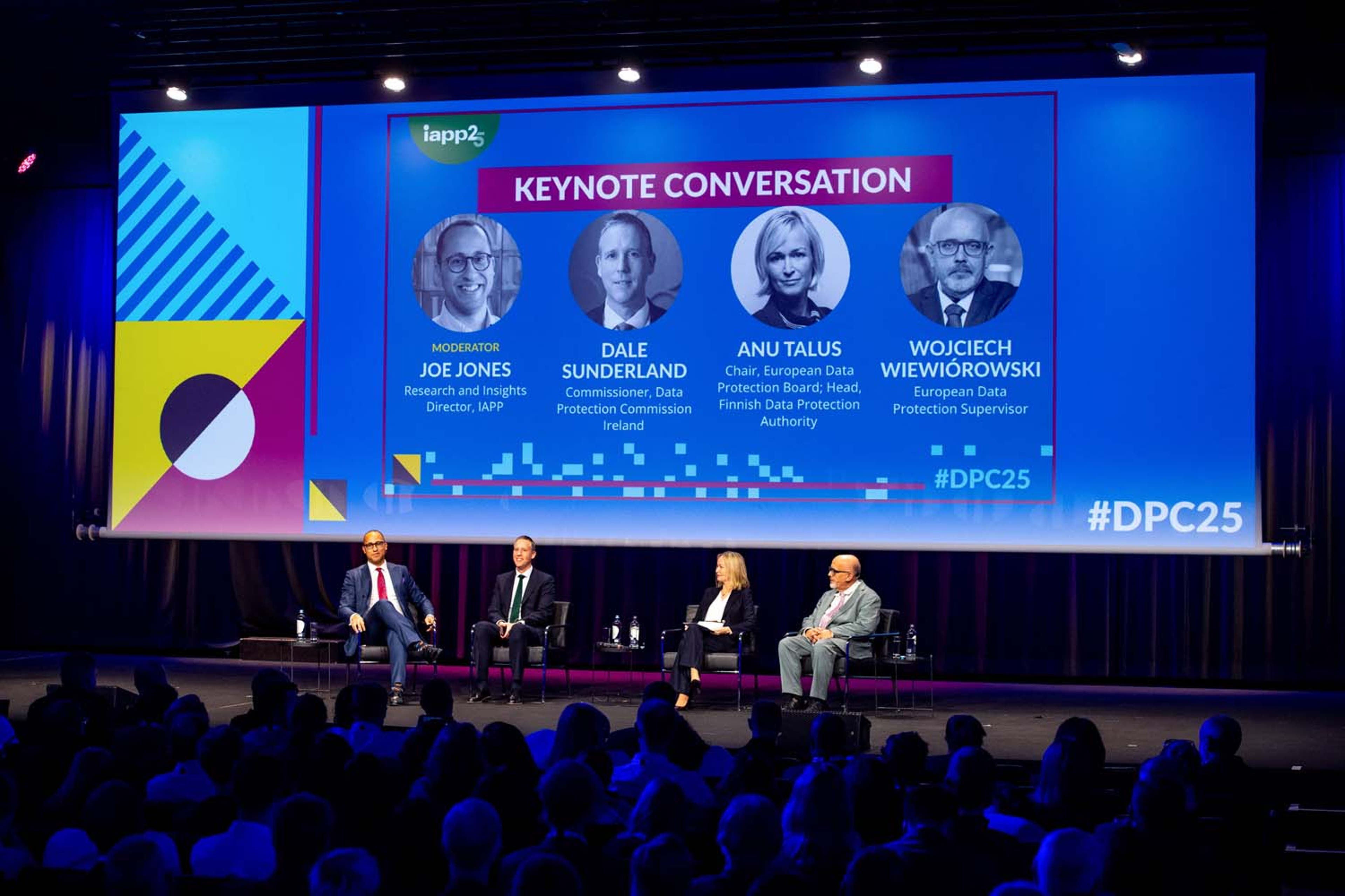 Panel discussion on stage with four speakers seated in front of a large screen displaying “Keynote Conversation” and names of participants.