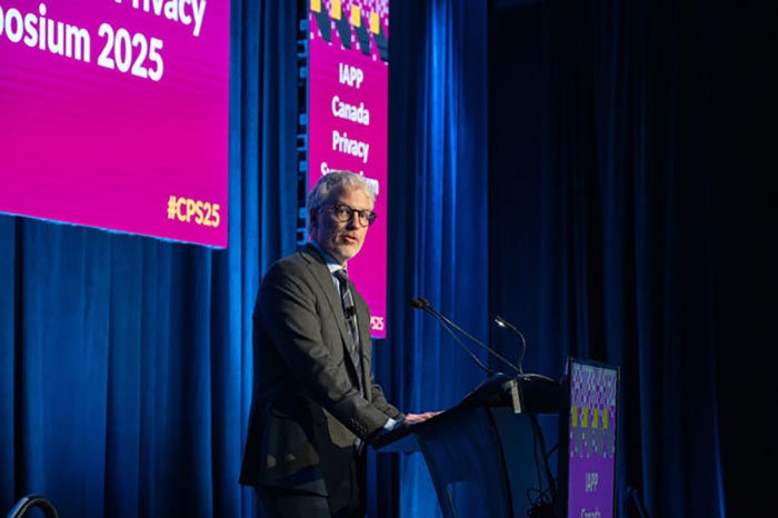 Speaker at a podium on stage with a blue curtain backdrop and pink signage reading “IAPP Canada Privacy Symposium 2025.”