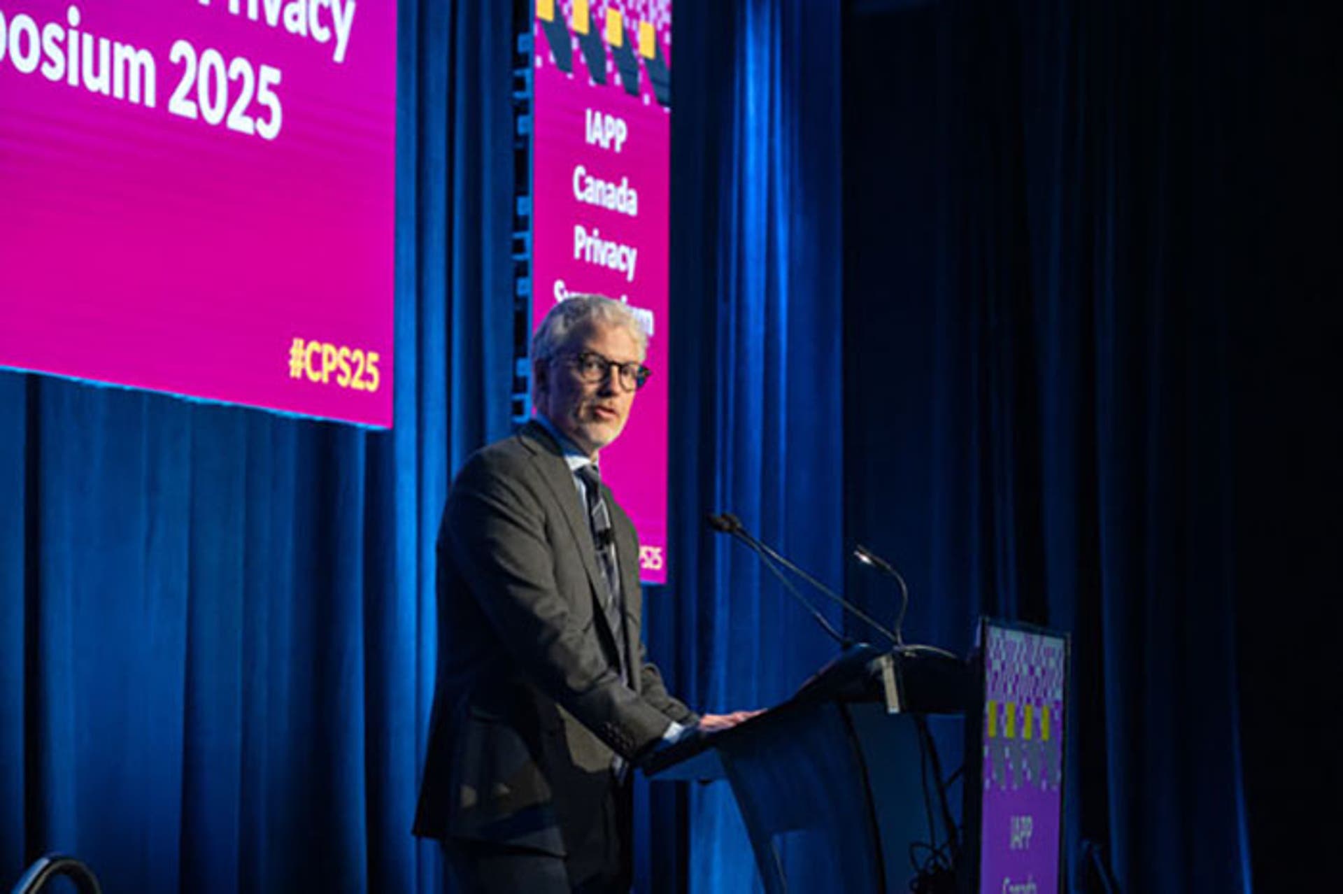 Speaker at a podium on stage with a blue curtain backdrop and pink signage reading “IAPP Canada Privacy Symposium 2025.”