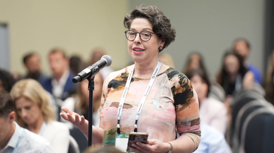 A woman with short hair and glasses stands at a microphone; she is asking a question during a workshop presentation.