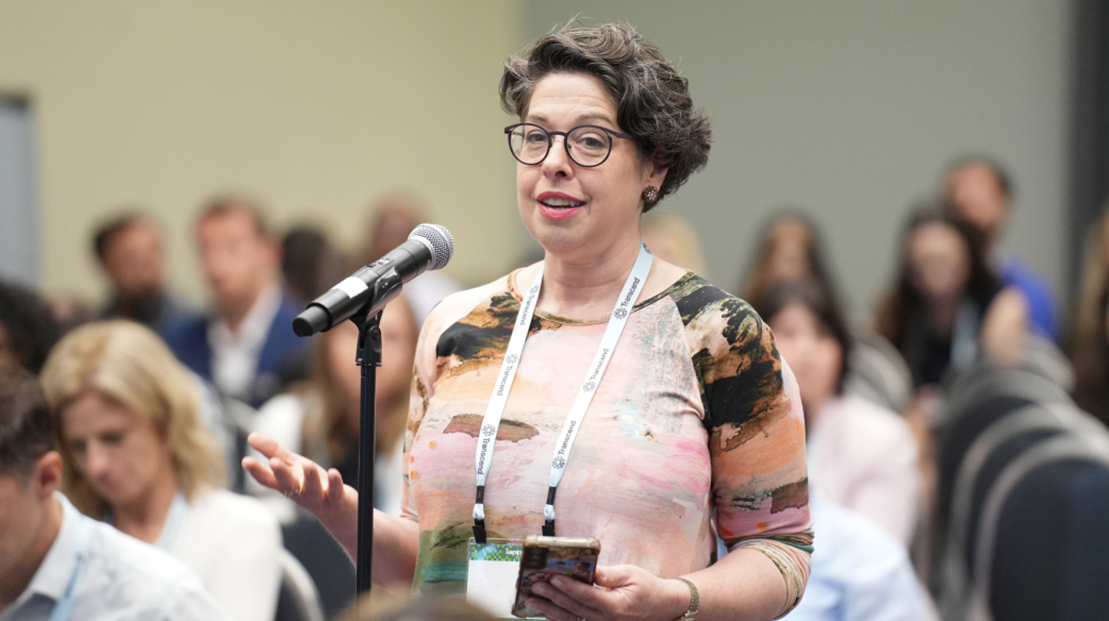 A woman with short hair and glasses stands at a microphone; she is asking a question during a workshop presentation.