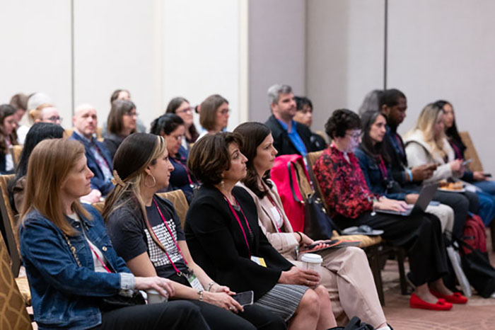 Audience seated in rows of chairs during a breakout session, some holding notebooks and laptops. A presentation screen is partially visible in the background.