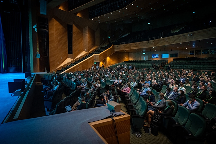 Large auditorium with rows of green seats filled with attendees listening to a presentation. Stage area is partially visible on the left.