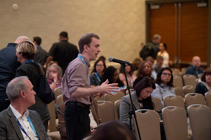 Attendee standing at a microphone in a conference room, speaking during a Q&A session. Rows of chairs and other participants are visible behind.