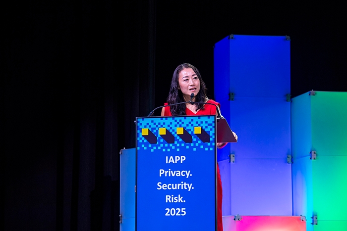 Speaker standing at a podium with a blue sign reading “IAPP Privacy. Security. Risk. 2025.” Brightly lit stage with stacked panels in blue, green, and purple hues behind.