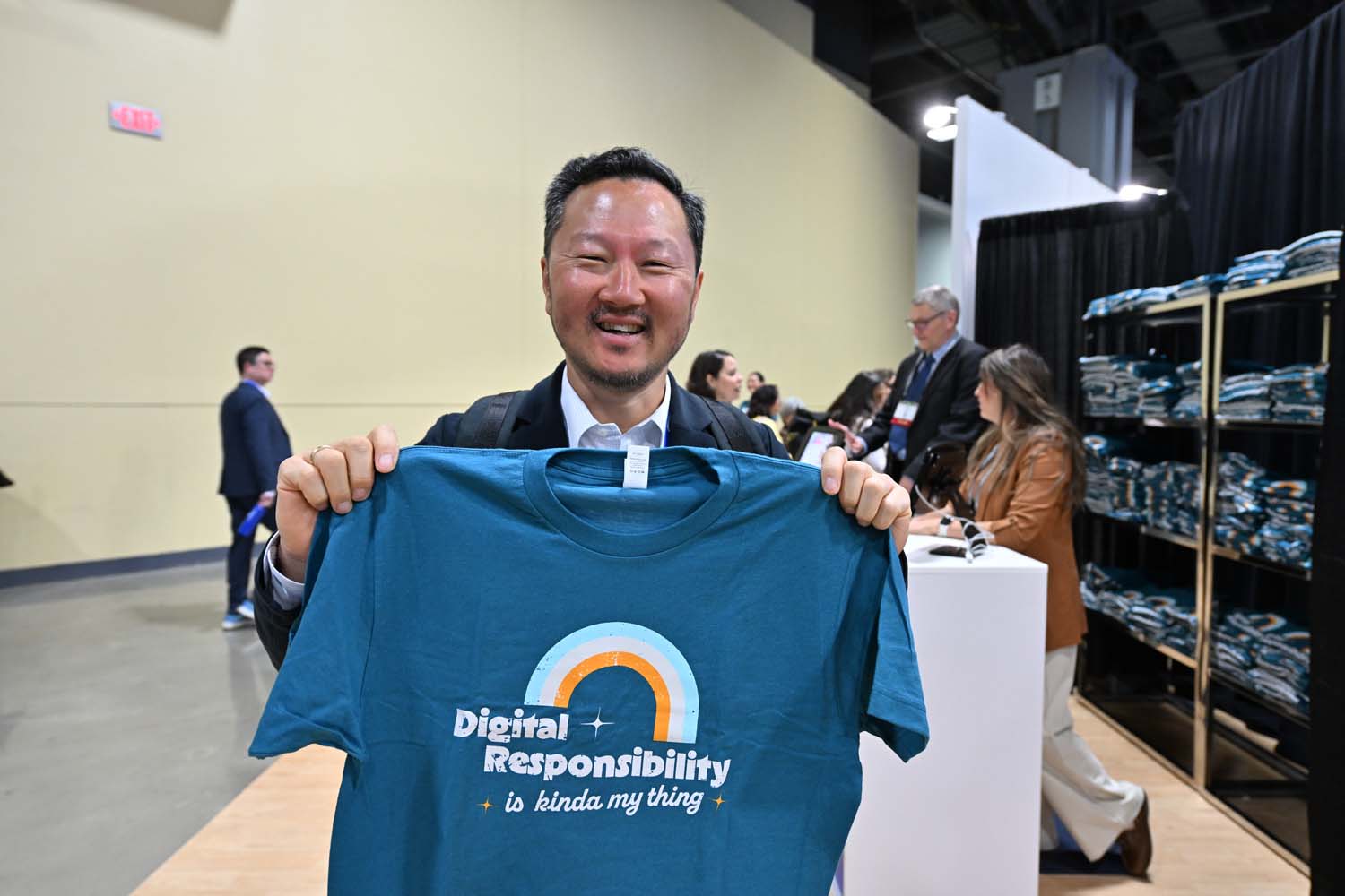 An attendee stands in a merchandise area holding up a teal T-shirt with the text Digital Responsibility is kinda my thing, with folded shirts and other conference participants visible in the background.