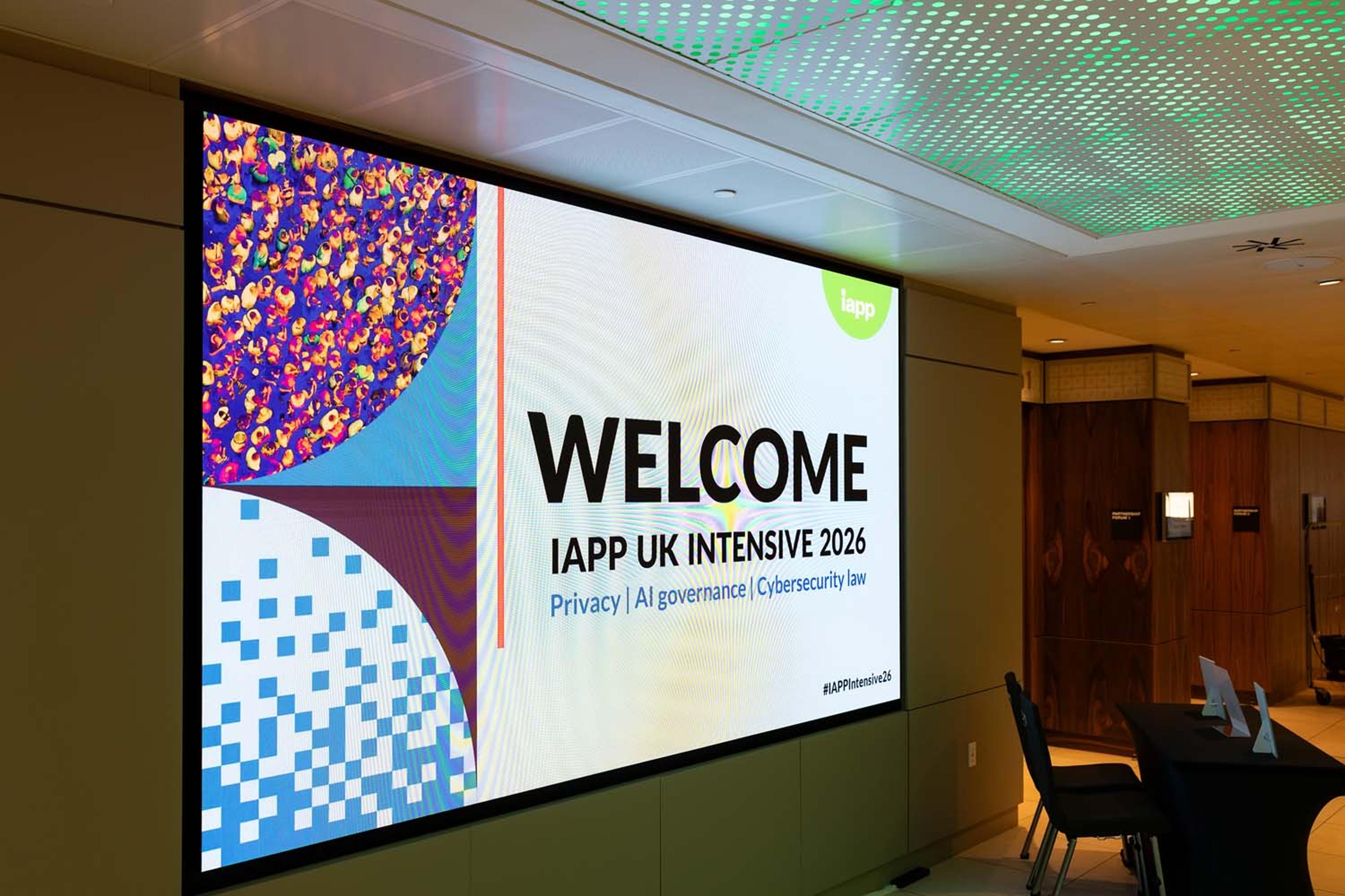 A large digital screen displays a welcome message for IAPP UK Intensive 2026 with privacy, AI governance, and cybersecurity law themes. Conference seating and tables are arranged nearby.