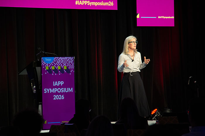 Presenter on stage at IAPP Symposium 2026 gesturing while speaking, with a branded podium displaying “IAPP SYMPOSIUM 2026” and a large pink event banner above.