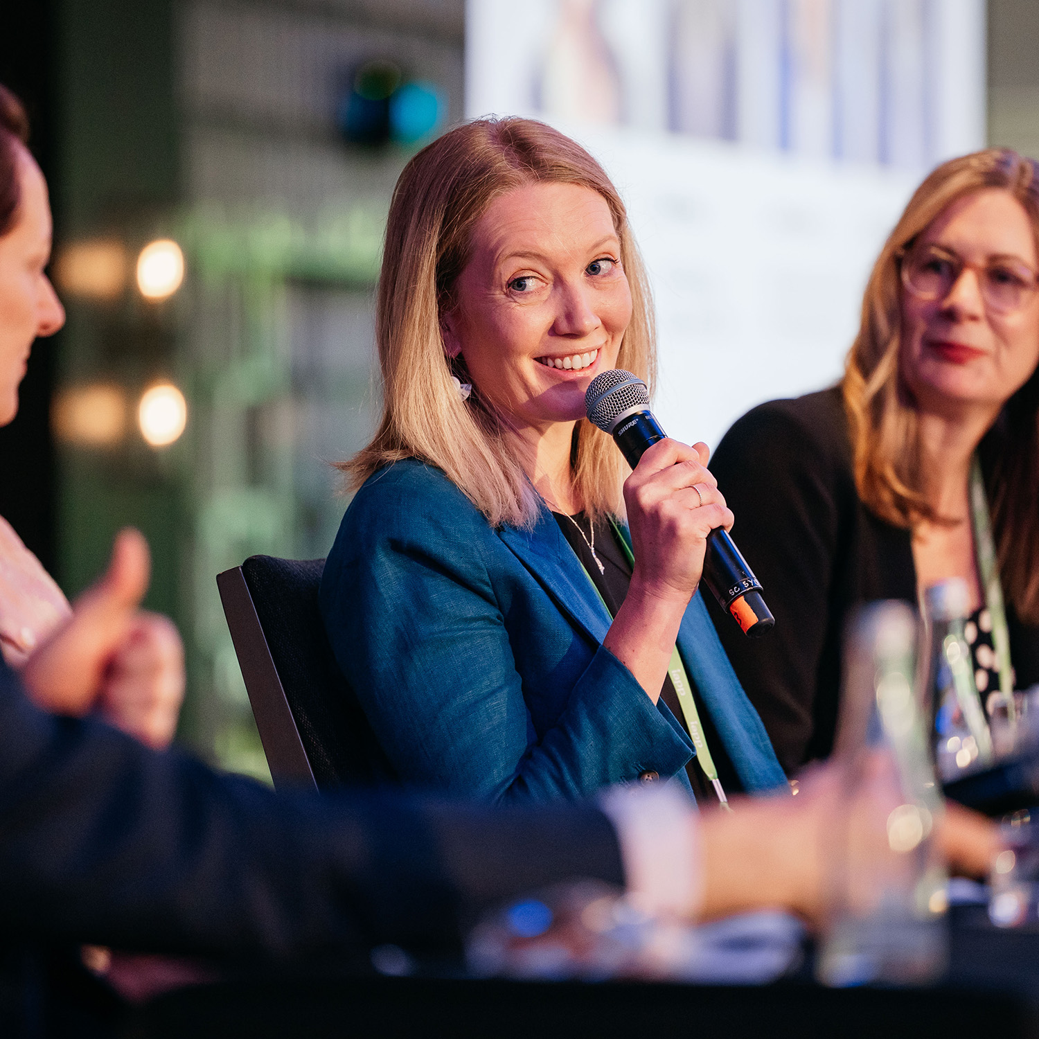 Close-up of a panel discussion with a person holding a microphone and speaking, seated at a table with water glasses.