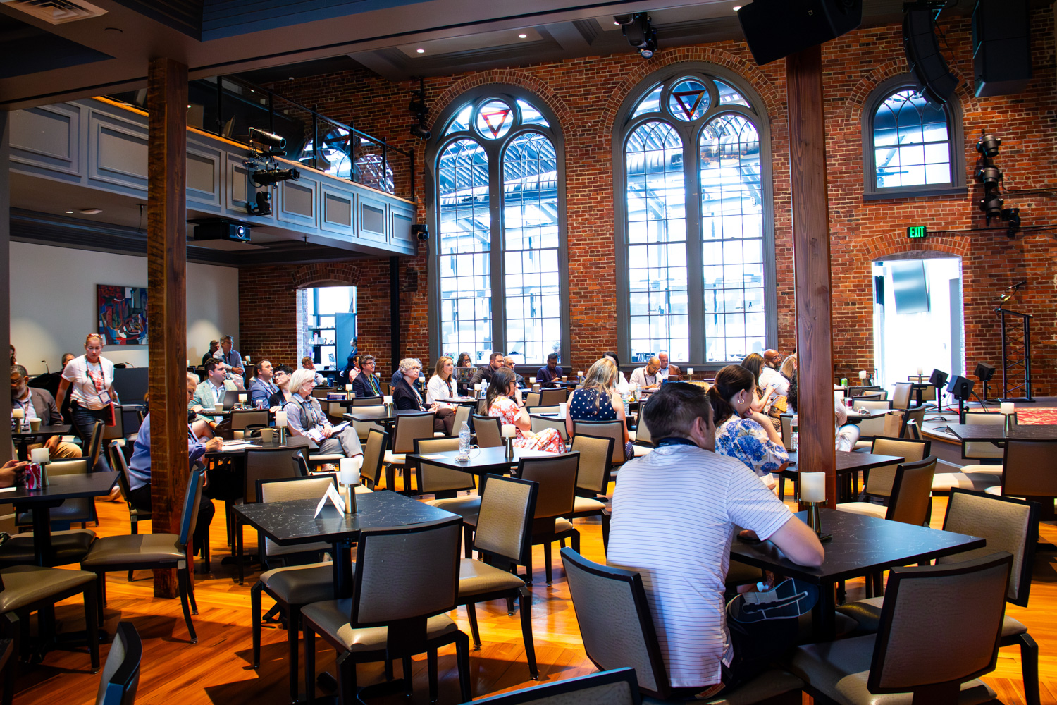 Attendees gather at tables inside a bright conference venue with exposed brick walls, tall arched windows, and polished wood floors. Participants face a presentation area at the front of the room, creating an atmosphere of attentive learning and discussion.