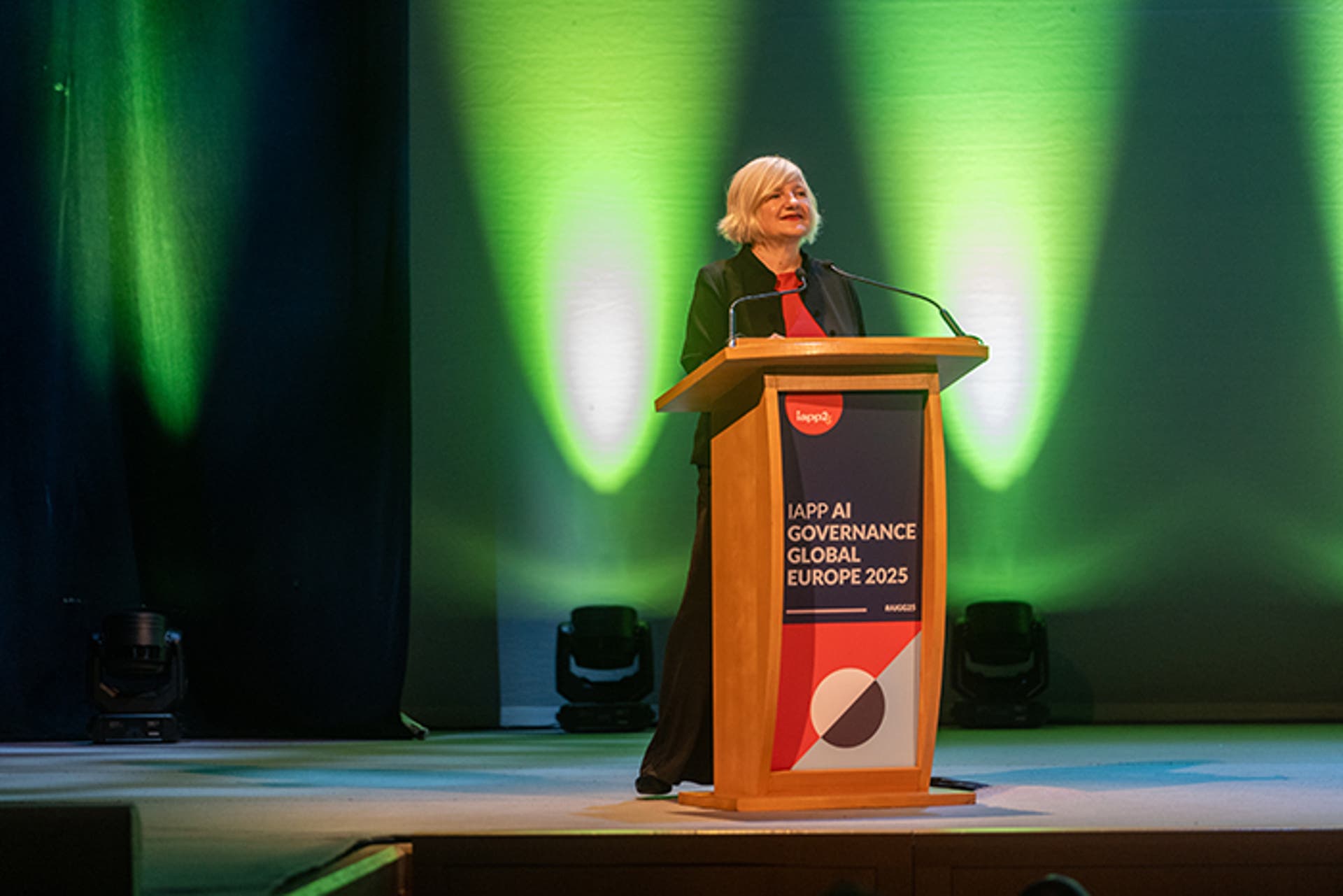 Speaker at a podium on stage with signage reading “IAPP AI Governance Global Europe 2025.” Green lighting illuminates the backdrop.