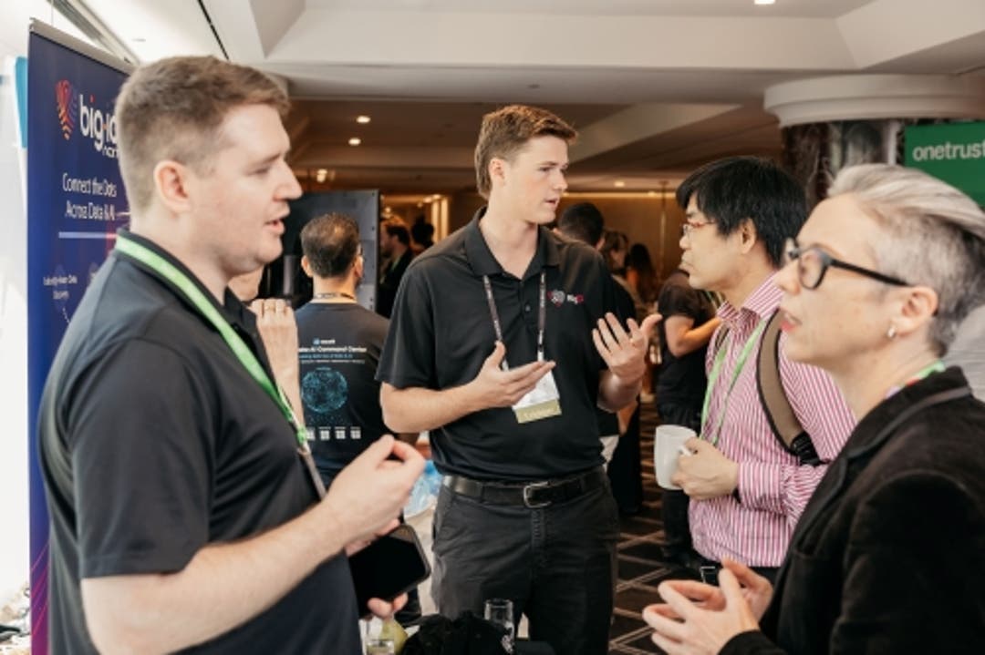Group of people networking at a conference near exhibitor booths with banners and lanyards visible.